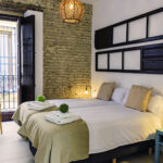 Dormitorio luminoso con dos camas individuales y acceso a balcón privado con vistas a la calle, destacando pared de ladrillo visto y decoración rústica en NintaiHome, Sevilla.