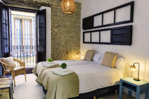 Dormitorio luminoso con dos camas individuales y acceso a balcón privado con vistas a la calle, destacando pared de ladrillo visto y decoración rústica en NintaiHome, Sevilla.