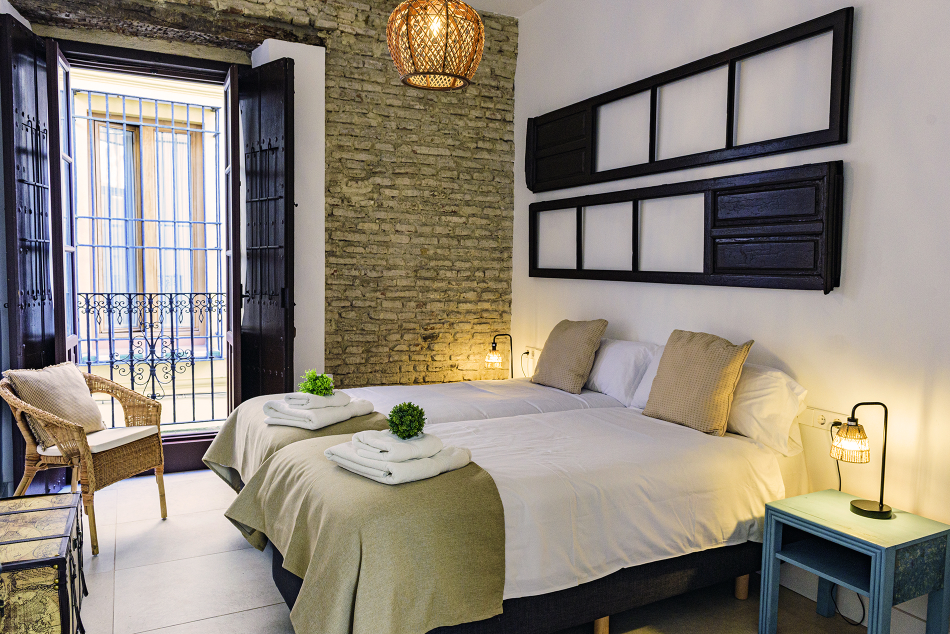 Dormitorio luminoso con dos camas individuales y acceso a balcón privado con vistas a la calle, destacando pared de ladrillo visto y decoración rústica en NintaiHome, Sevilla.